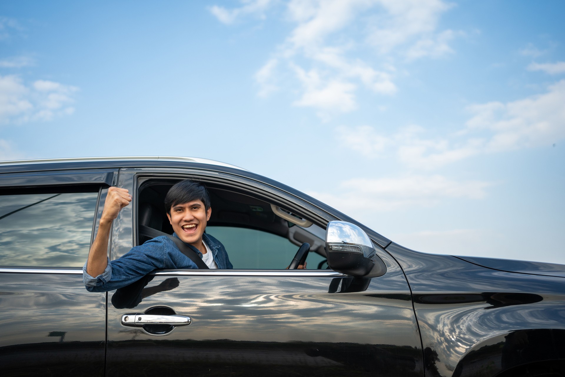 A young Asian man drives a car on a clear day. With beautiful blue sky. He smiling driving to travel by car. Sticking her head outta the windshield
